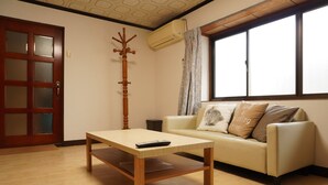 A living room with a wooden table, a sofa with cushions, a coat rack, and a window with curtains. - Private House for Rabbit Island (Takehara)