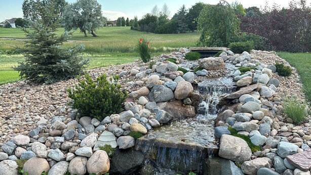 Ein Garten mit einem kleinen Wasserfall, umgeben von Steinen und viel Grün.