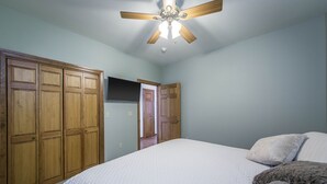 A bedroom with a bed, wooden wardrobe, ceiling fan, and a door leading to another room.