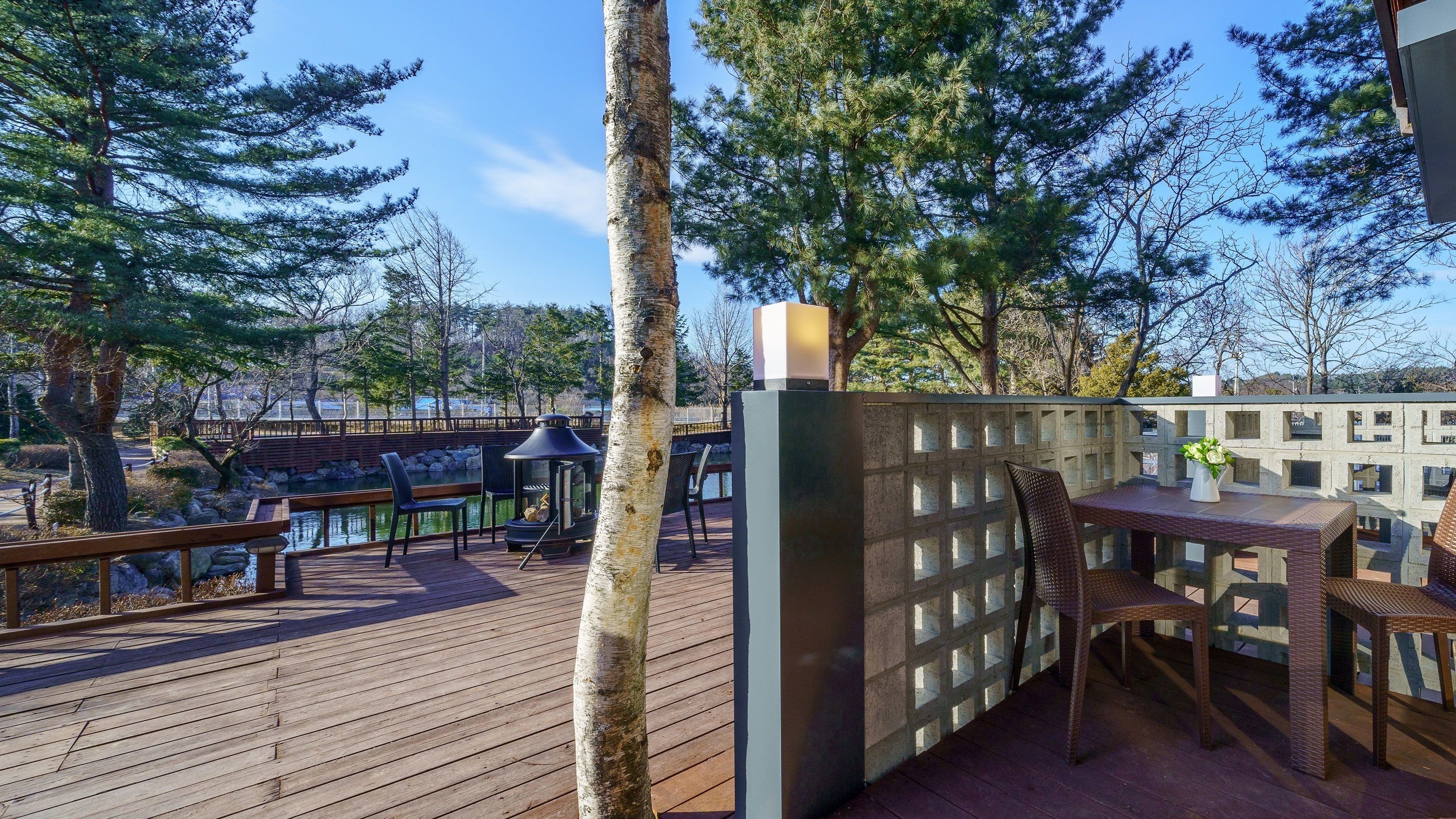 Eine Holzterrasse mit Sitzgelegenheiten im Freien, einer Feuerstelle und Blick auf ein Gewässer und Bäume.