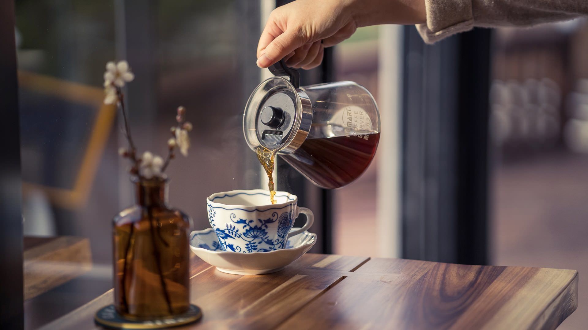 A hand pouring tea from a glass teapot into a blue and white cup on a wooden table.