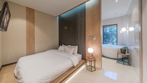 A modern bedroom with a bed, a bedside table with a lamp, a window with a view of trees, and a bathroom with a bathtub and a plant.
