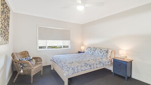 Une chambre à coucher comprenant un lit, deux tables de chevet, une chaise, une fenêtre avec des stores et un ventilateur de plafond.