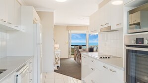 Une cuisine moderne avec des meubles blancs, un four encastré et une vue sur l’océan grâce à une porte-fenêtre coulissante.