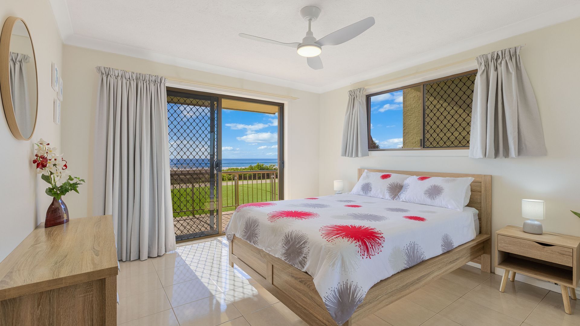 Une chambre à coucher comprenant un lit, une table de chevet, un bureau, un vase avec des fleurs et une vue sur l’océan depuis un balcon.
