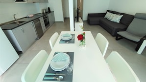 Una cocina moderna con microondas, horno y pileta. Hay una mesa de comedor con platos, cubiertos y un florero con una rosa roja. Un sofá gris y un cuadro con una playa se encuentran en la sala de estar.