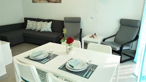 Un comedor con una mesa blanca para dos, un sofá gris, un sillón gris y una pequeña mesa blanca con una lámpara.