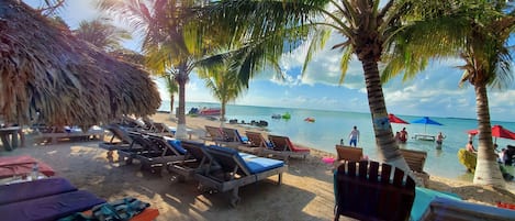 A beach with lounge chairs, palm trees, and a thatched roof structure.