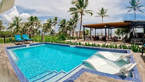A swimming pool with blue-tiled edges, lounge chairs, and palm trees in the background. - Mangata Villas (San Pedro)