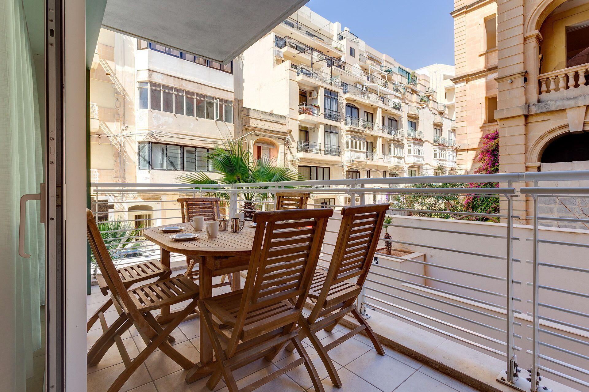 A balcony with a wooden table and chairs, overlooking a row of multi-story buildings with balconies.