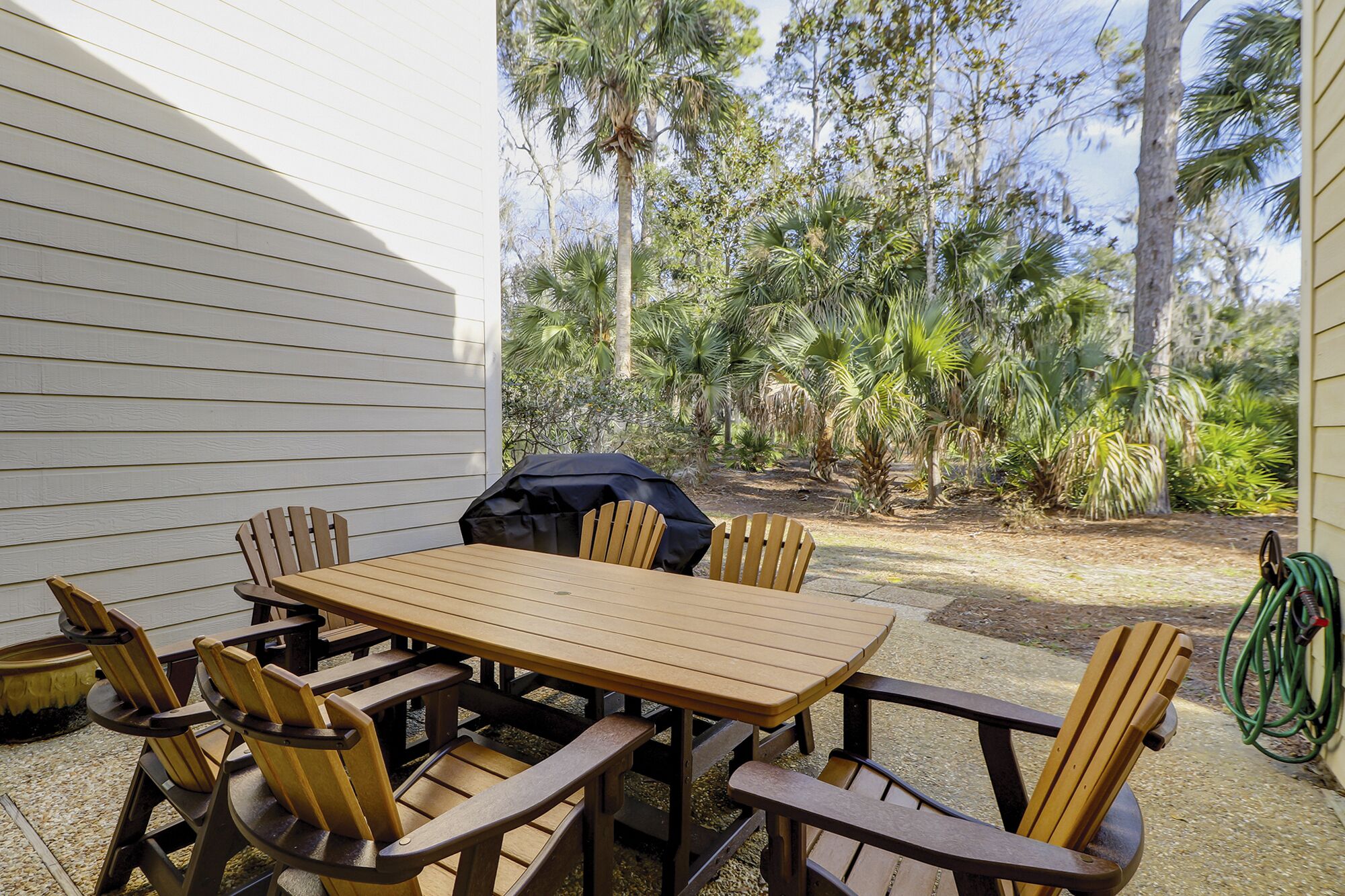 Een houten tafel met stoelen buiten, met een barbecue en groen in de achtergrond.