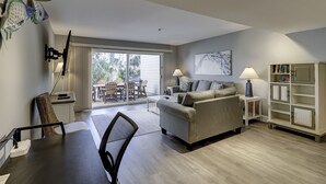 A modern living room with a sofa, a dining table, and a TV. There's a sliding glass door leading to a patio with outdoor furniture. - Sunset Rentals | Shipmaster 1602 | Dog Friendly (Hilton Head Island)