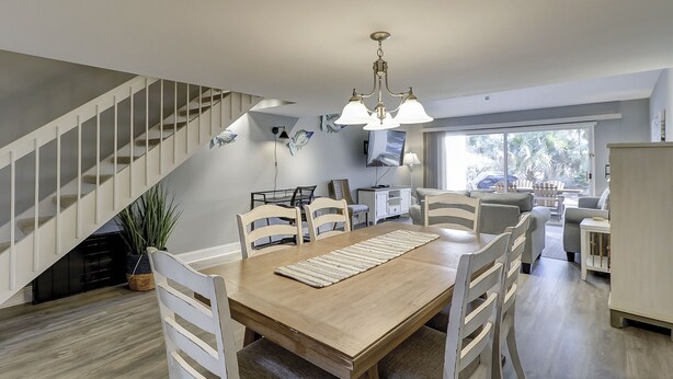 A spacious dining area with a wooden table and white chairs, a staircase, and a large window with a view of greenery. - Sunset Rentals | Shipmaster 1602 | Dog Friendly (Hilton Head Island)