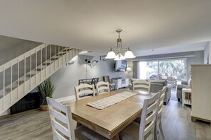 Un comedor amplio con una mesa de madera y sillas blancas, una escalera y un gran ventanal con vistas al verdor.
