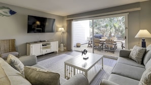 A living room with a TV, white entertainment unit, and a sliding glass door leading to an outdoor patio with chairs and a table. - Sunset Rentals | Shipmaster 1602 | Dog Friendly (Hilton Head Island)