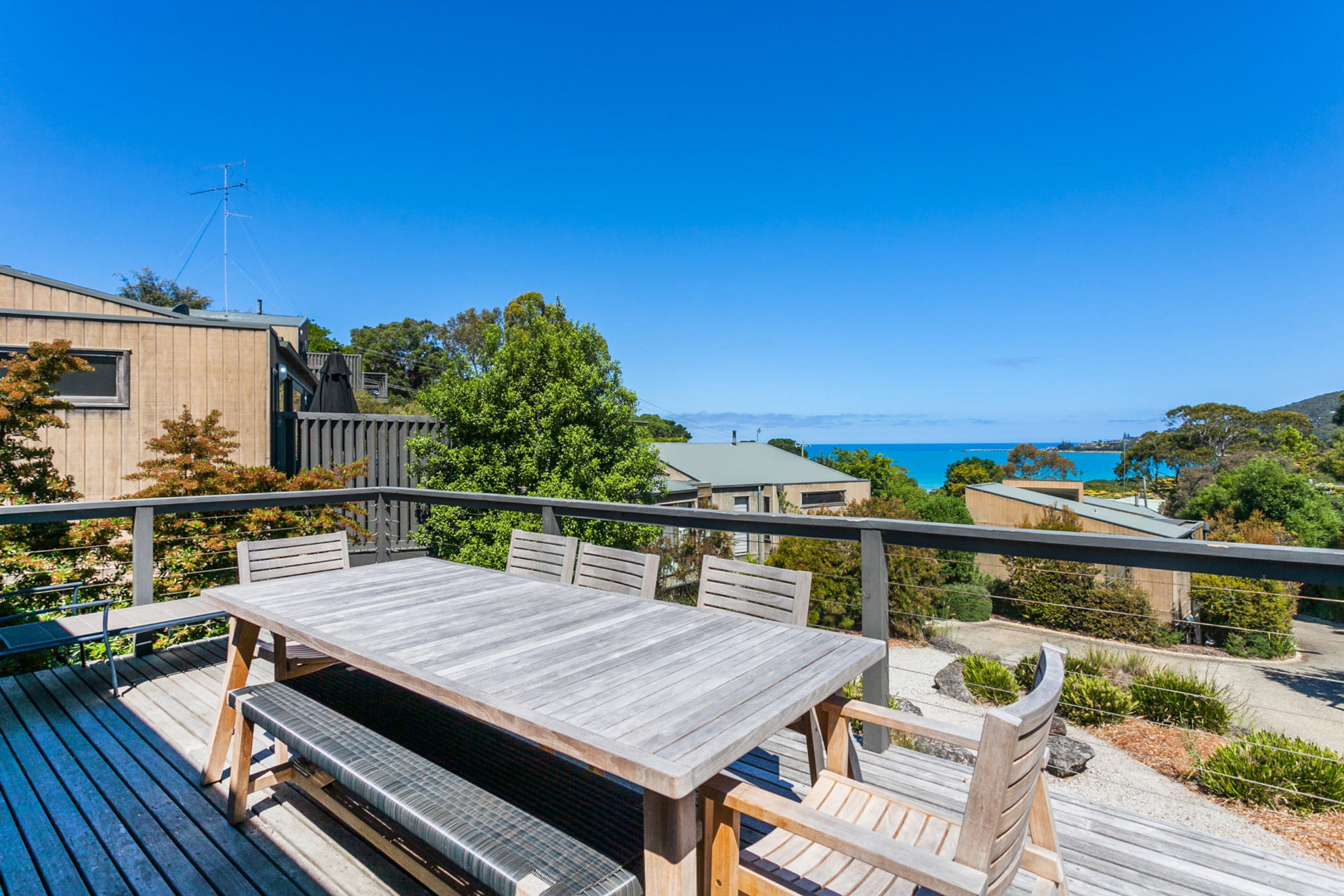A wooden deck with a table and chairs, overlooking a coastal view with buildings and trees.