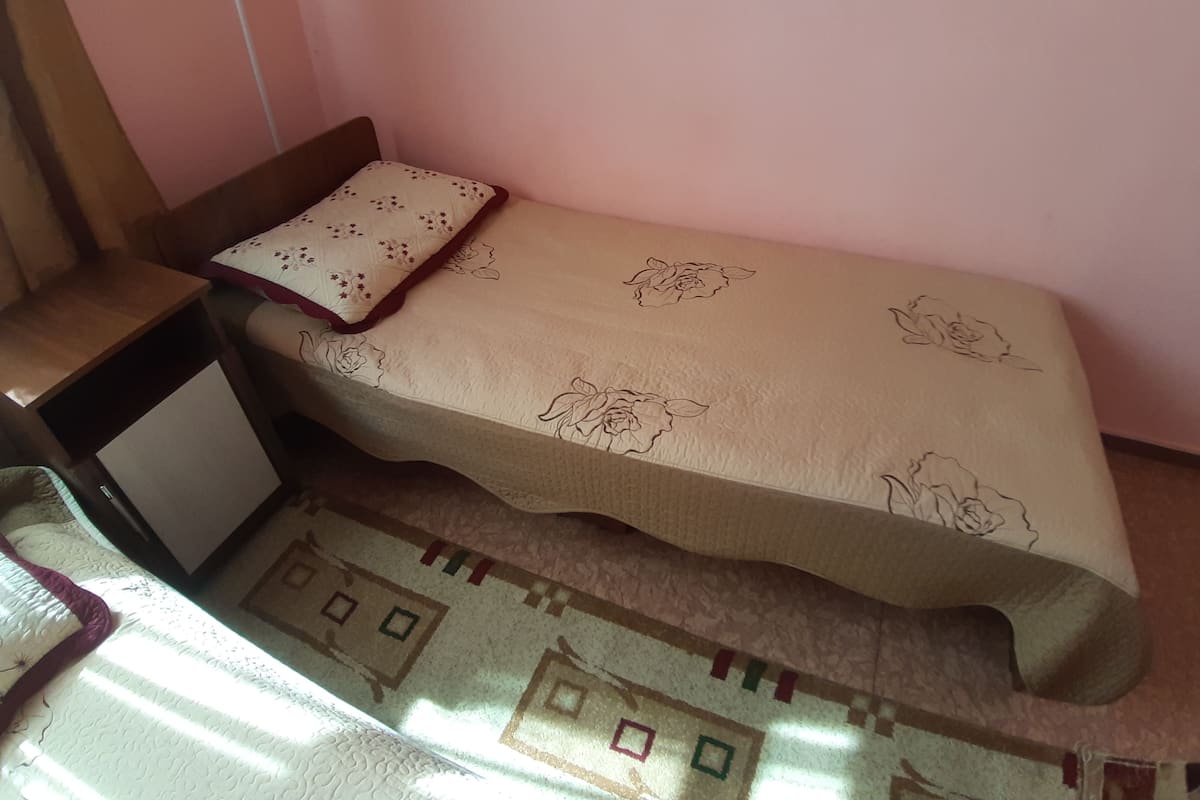 Une petite chambre avec un lit simple, une table de chevet en bois et une couverture à motifs.