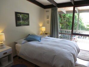 A bedroom with a bed, a nightstand, a lamp, a picture on the wall, and a window with a view of greenery. - Beachview (Lorne)
