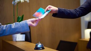 A hand holding a colorful card, possibly a business card, being handed over at a reception desk with a bell and a plant in the background.