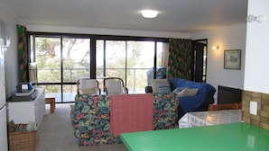 A room with a green table, two chairs, a sofa with floral cushions, a window with curtains, and a small table with a radio.