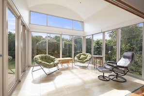 Ein geräumiges Zimmer mit großen Glasfenstertüren, die zu einem Garten führen, mit zwei grünen Stühlen und einem braunen Ledersessel.
