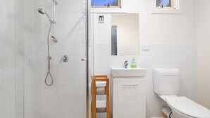 A bathroom with a shower, toilet, sink, and a small wooden stool. - Coastal Haven (Anglesea)