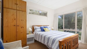 A bedroom with a large wooden wardrobe, a single bed with striped bedding, a nightstand, and a window with curtains. - Coastal Haven (Anglesea)