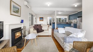 A living room with a fireplace, a sofa, a coffee table, and a kitchen in the background. - Coastal Haven (Anglesea)