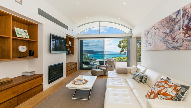 A modern living room with a large TV, wooden shelves, a sofa, and a view of the ocean. - The Pavillion (Lorne)