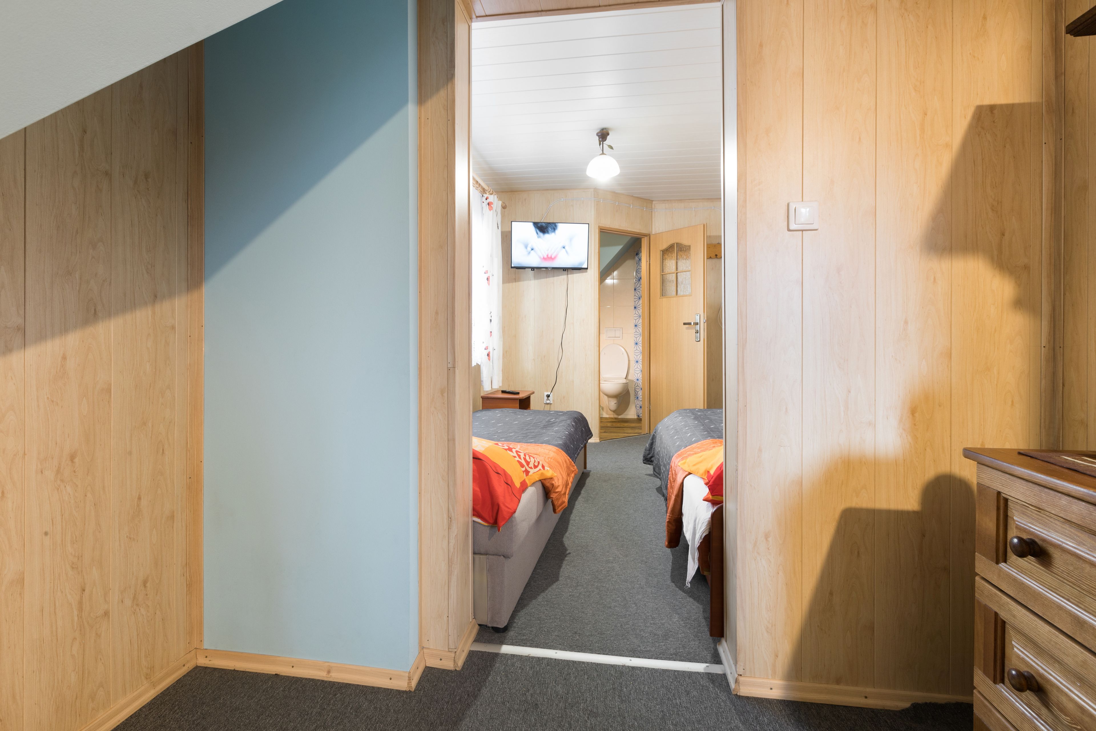 A narrow hallway with wooden walls and carpeted floors leading to two bedrooms with beds and a television.