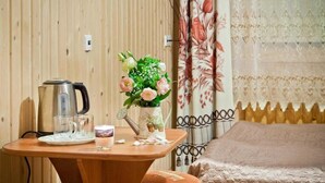 Una mesa de madera con un hervidor, vasos y un arreglo floral. Una cama con cabecera estampada se ve al fondo.