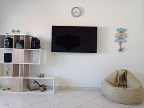 A living room with a flat-screen TV mounted on the wall, a white shelf unit with various items, a beige bean bag chair, and a wall clock. - Gaveto- Sea Front, Refurbished and Cozy (Quarteira)