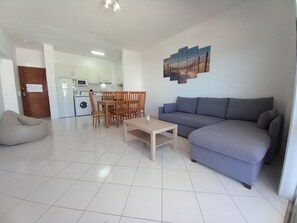 A modern living room with a grey sofa, wooden dining table, and chairs. A beige bean bag is on the floor. A kitchen with white appliances is visible in the background.