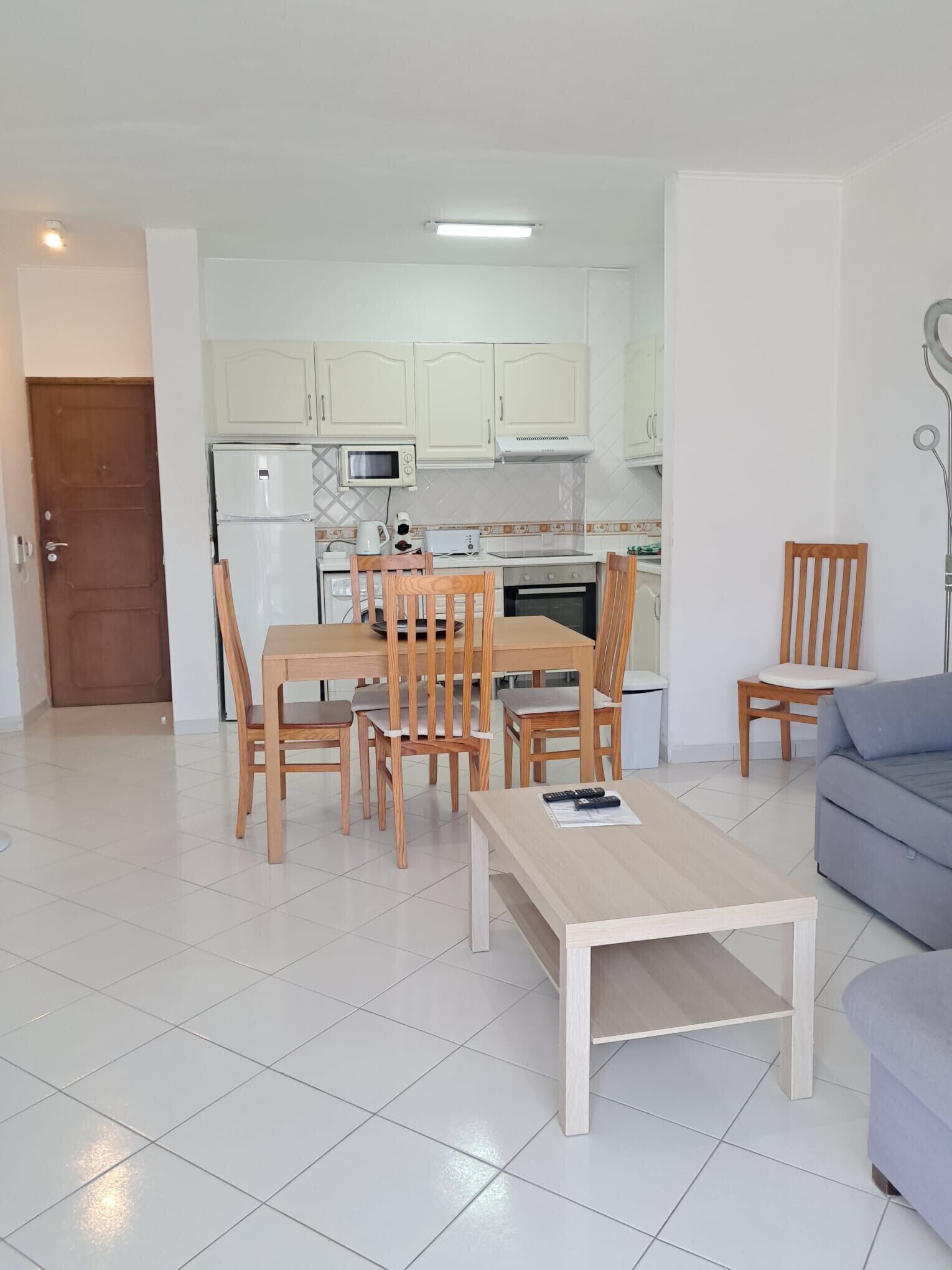 A modern kitchen with white cabinets, a dining table with chairs, and a sofa in a living area with tiled flooring.
