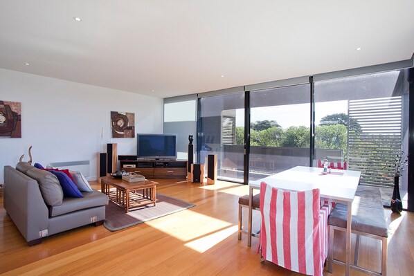 A modern living room with a flat-screen TV, a sofa, a dining table with chairs, and a large sliding glass door leading to an outdoor area.