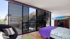 A modern bedroom with a large sliding glass door, a bed with a purple blanket, a television, and a painting of a beach scene. - Debonair 1 (Anglesea)