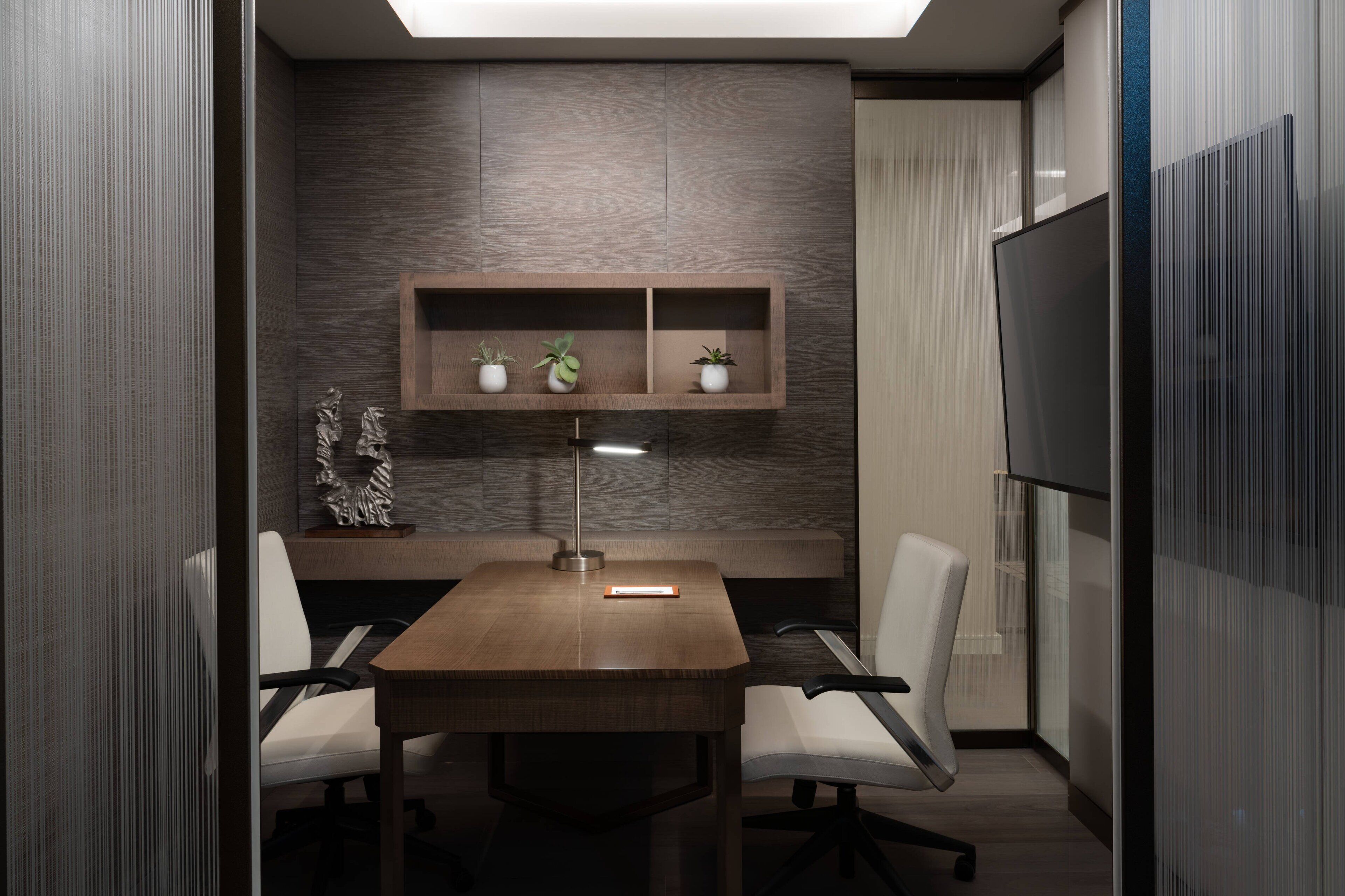 Een vergaderruimte met een houten tafel, twee witte stoelen, een bureaulamp en een plank met potplanten.
