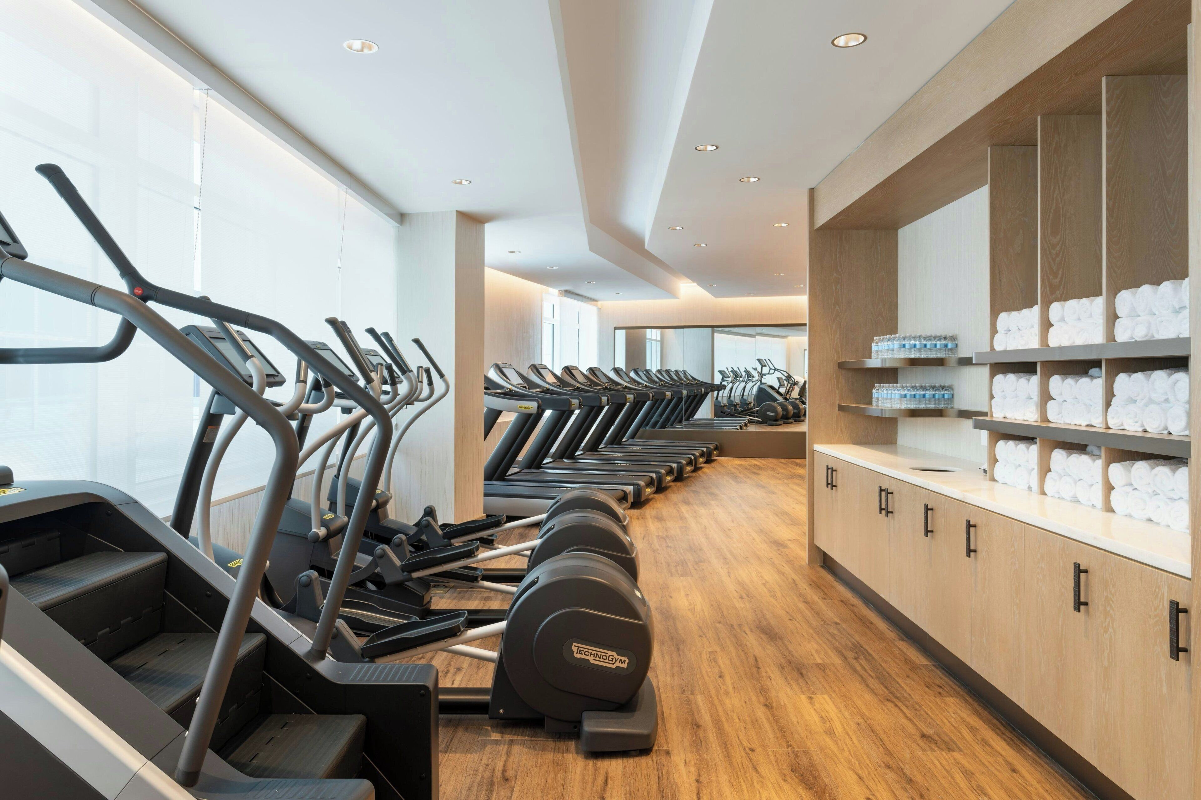 Un gimnasio con varias cintas de correr alineadas, suelo de madera y una pared con estantes que contienen toallas.