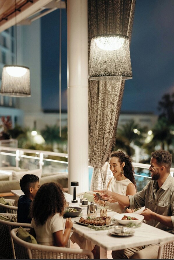Comedor con mesa lista para cuatro personas, rodeado de sillas de mimbre, bajo lámparas colgantes.