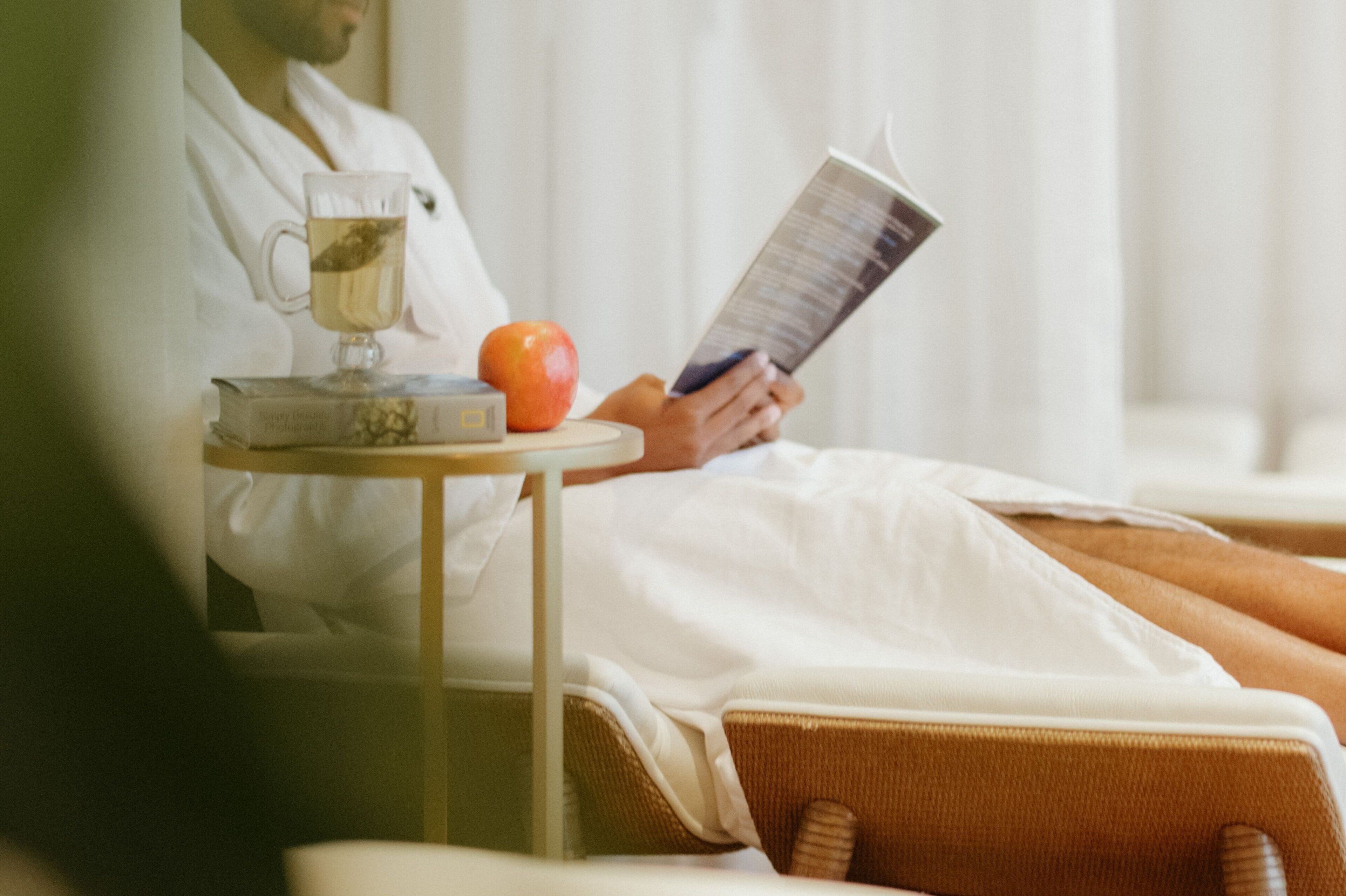 A person reclining in a spa chair, reading a book, with a glass of tea and an apple on a small table beside them.