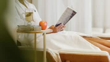 A person reclining in a spa chair, reading a book, with a glass of tea and an apple on a small table beside them.
