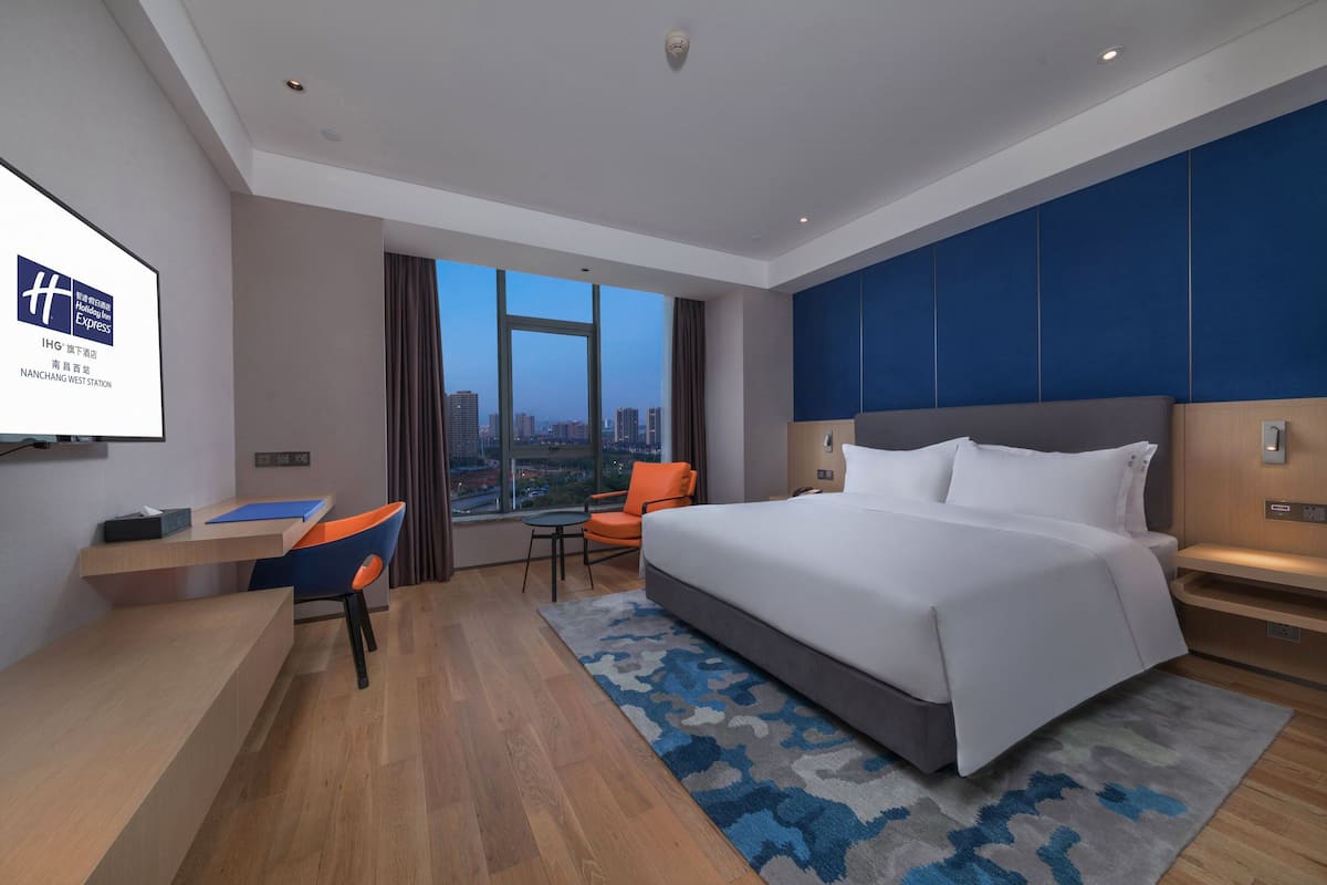 Une chambre d’hôtel moderne avec un grand lit, un bureau et une chaise. Une grande fenêtre offre une vue sur la ville.