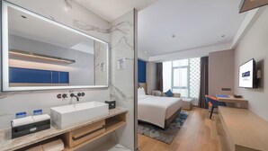 Una habitación de hotel moderna con un espejo grande, un lavamanos blanco, una cama, un escritorio y una televisión.