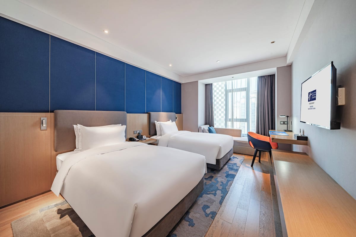 Une chambre d’hôtel avec deux lits, un bureau, une chaise, une télévision et une grande fenêtre avec des rideaux.