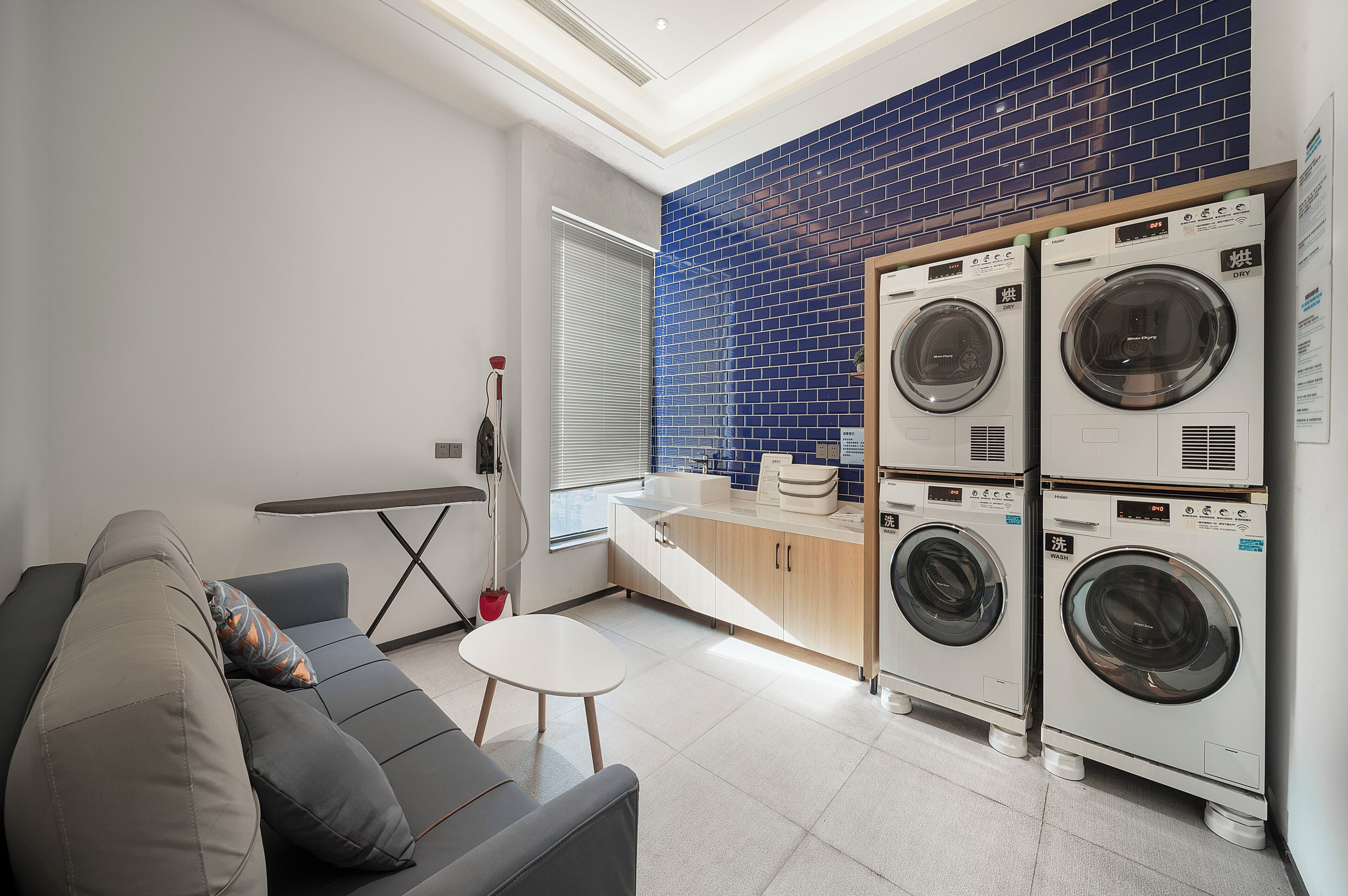 A compact laundry room with two stacked washing machines, a grey sofa with cushions, a small white table, and a window with blinds.