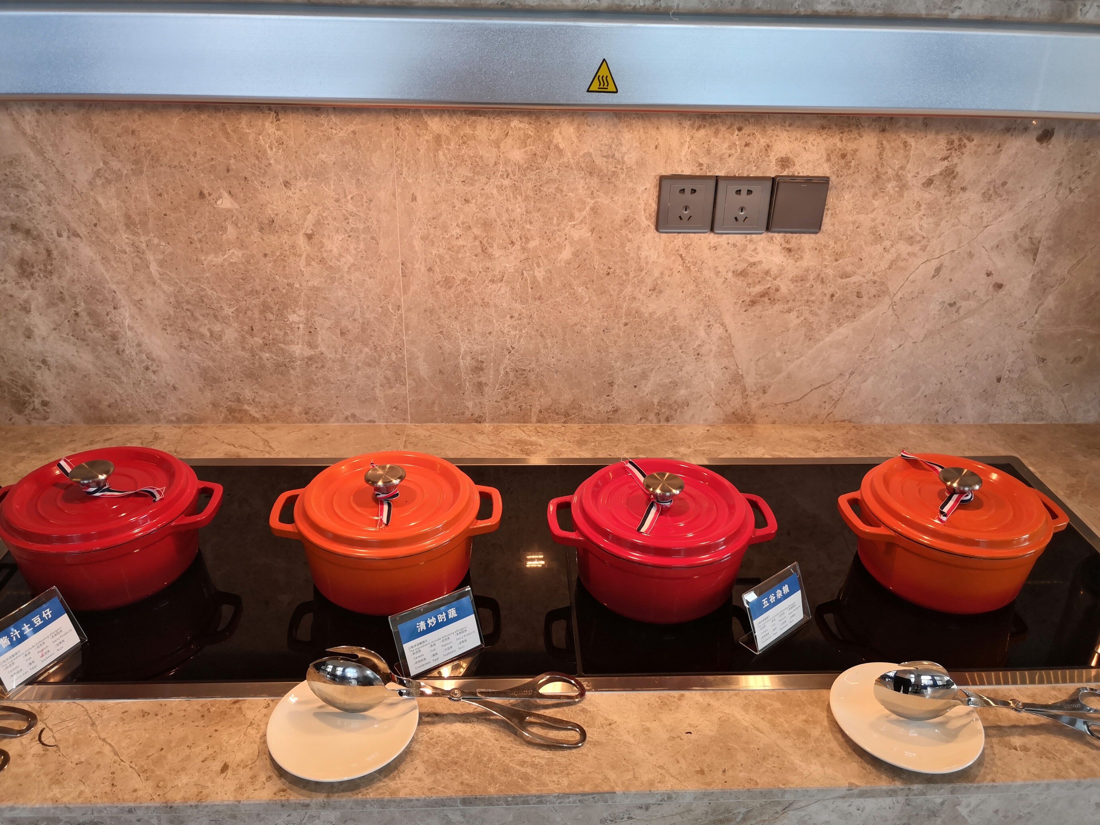 Four red pots on a stove with handles, a warning sign above electrical outlets, and a metal spoon on a plate.