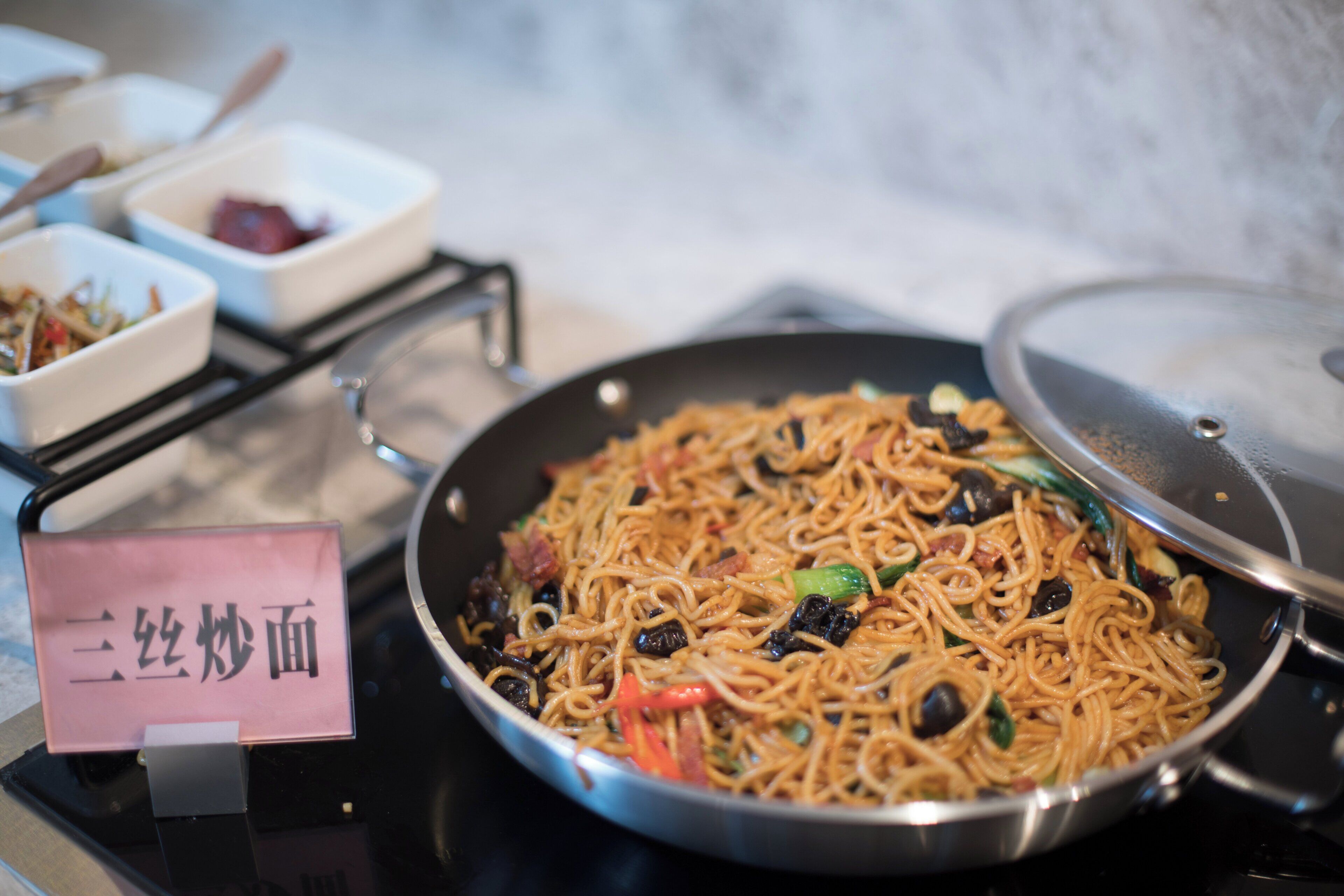 Una sartén de fideos salteados con verduras y carne, un tazón pequeño de salsa roja y un letrero con caracteres chinos.