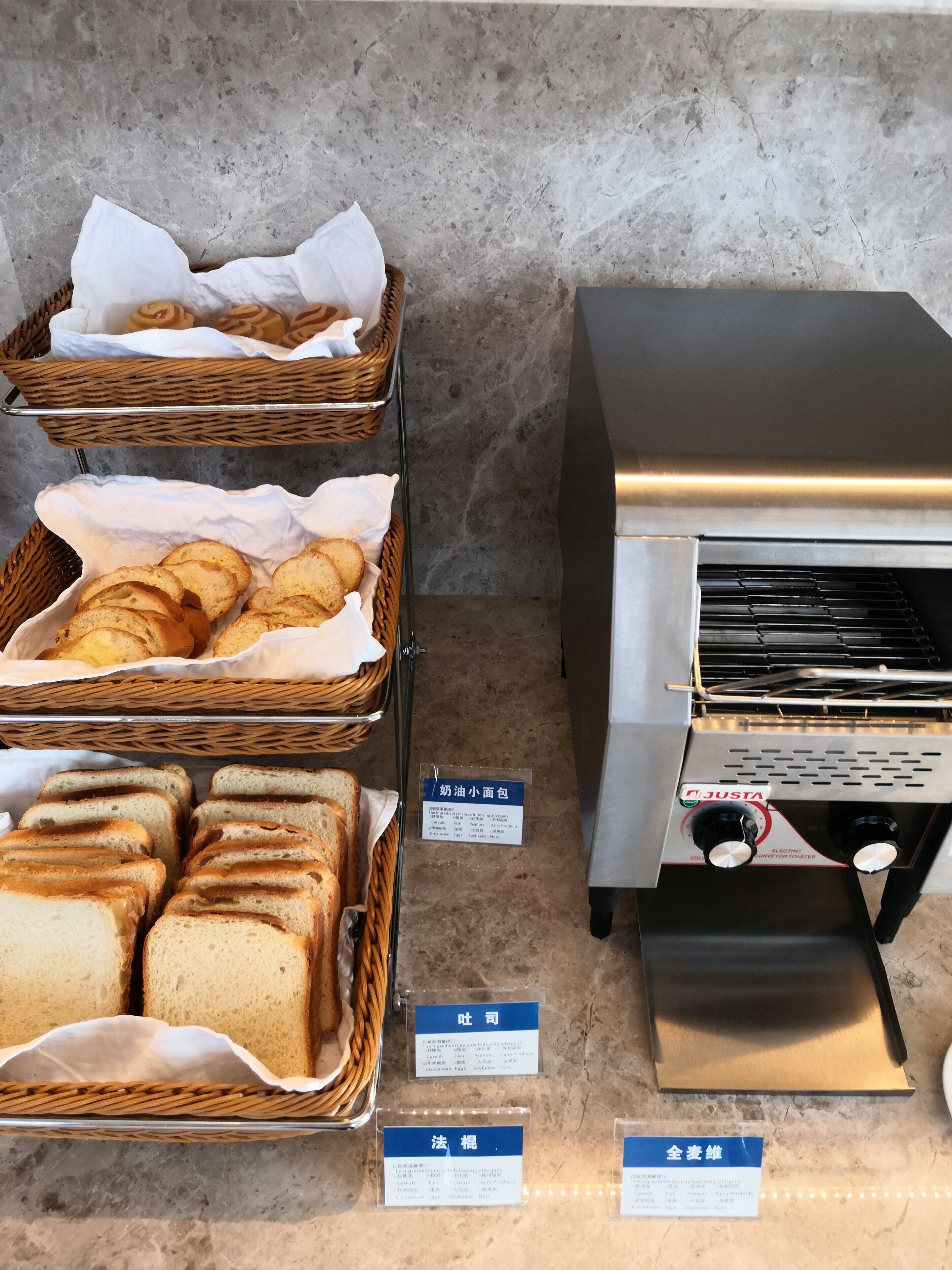 Un expositor de panadería con panes y pasteles en cestas de mimbre, un tostador industrial y etiquetas que indican los tipos de productos.