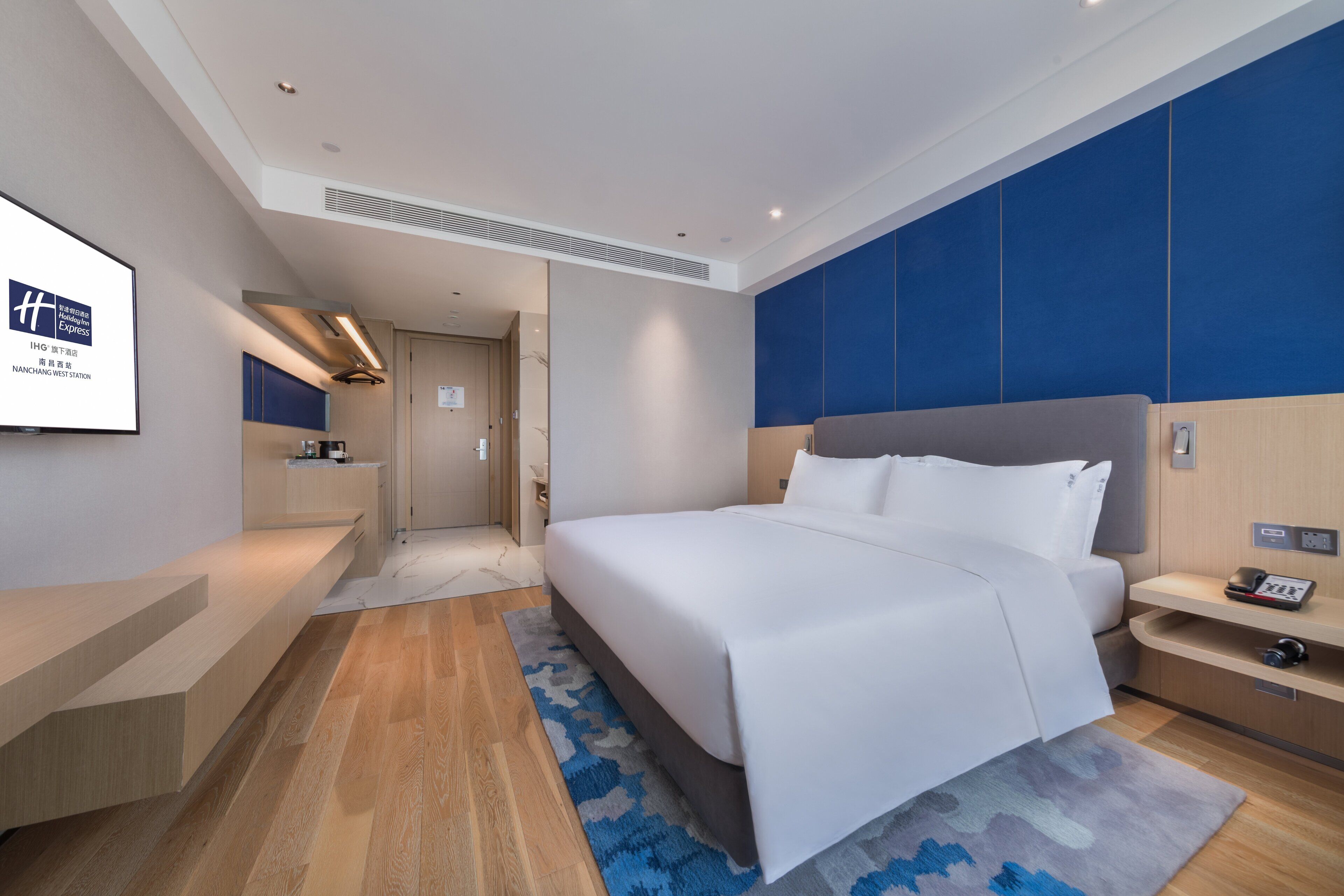 A modern hotel room with a large bed, a bedside table, a built-in TV, and a wooden floor.