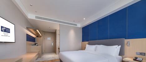 A modern hotel room with a large bed, a bedside table, a built-in TV, and a wooden floor.